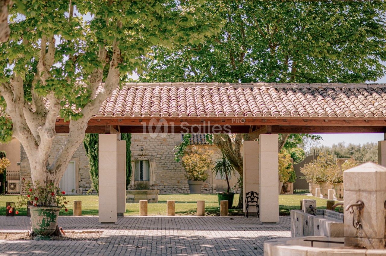 Cour ombragée avec toit en tuiles, arbres, bancs en pierre et végétation devant un bâtiment en pierre.
