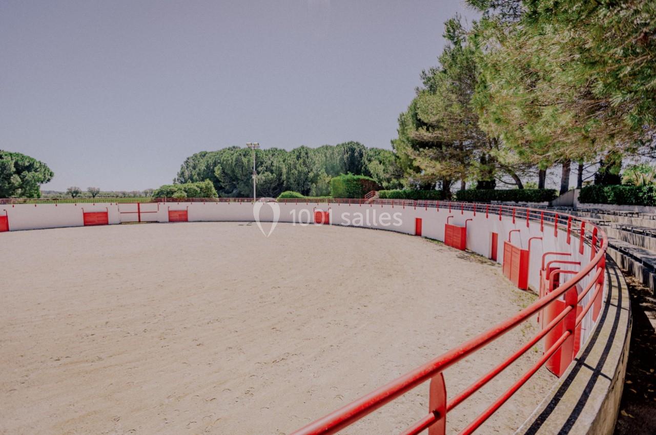 Arène circulaire en plein air avec sol en sable, entourée de barrières rouges et blanches, bordée d'arbres.