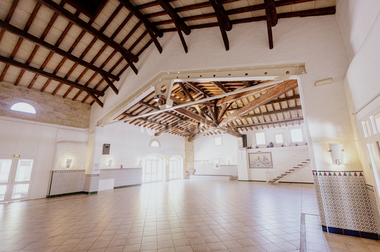 Grande salle lumineuse avec plafond en poutres apparentes, murs blancs et carrelage au sol, éclairée par des fenêtres.