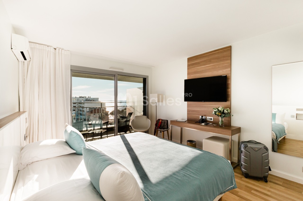 Chambre d'hôtel lumineuse avec lit double, balcon donnant sur la ville, télévision murale et valise posée au sol.