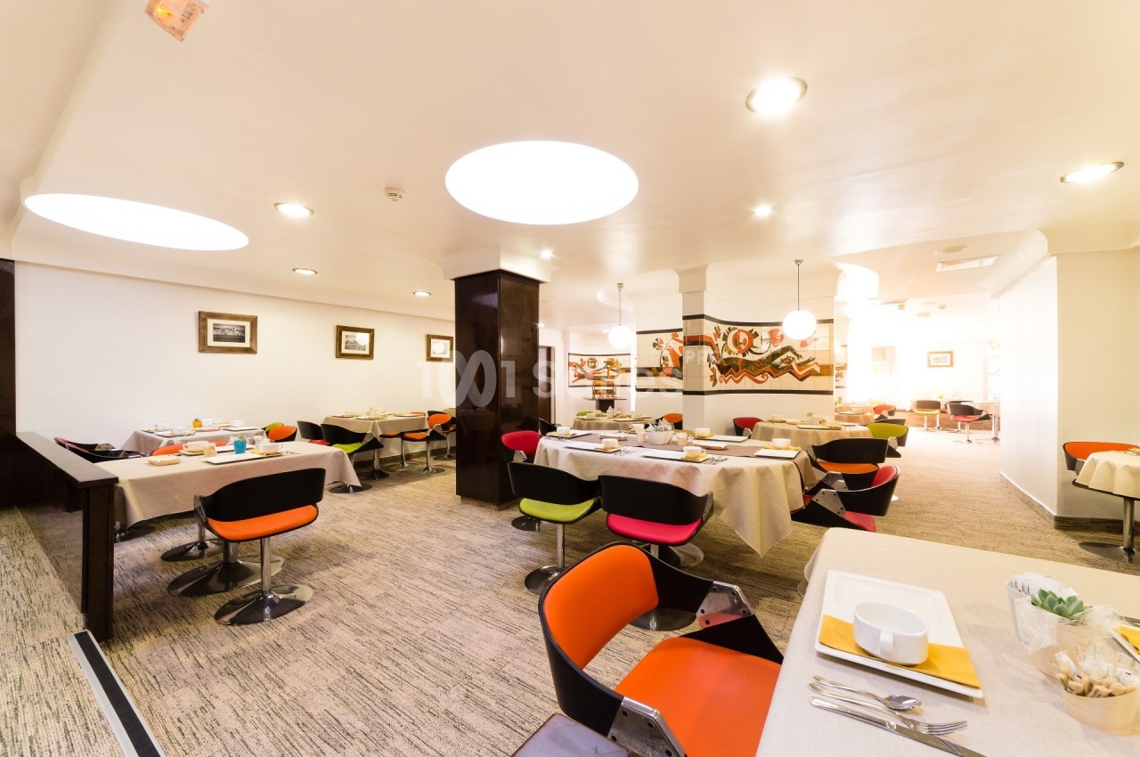 Salle lumineuse avec tables dressées pour le petit-déjeuner, chaises colorées et décor mural abstrait.