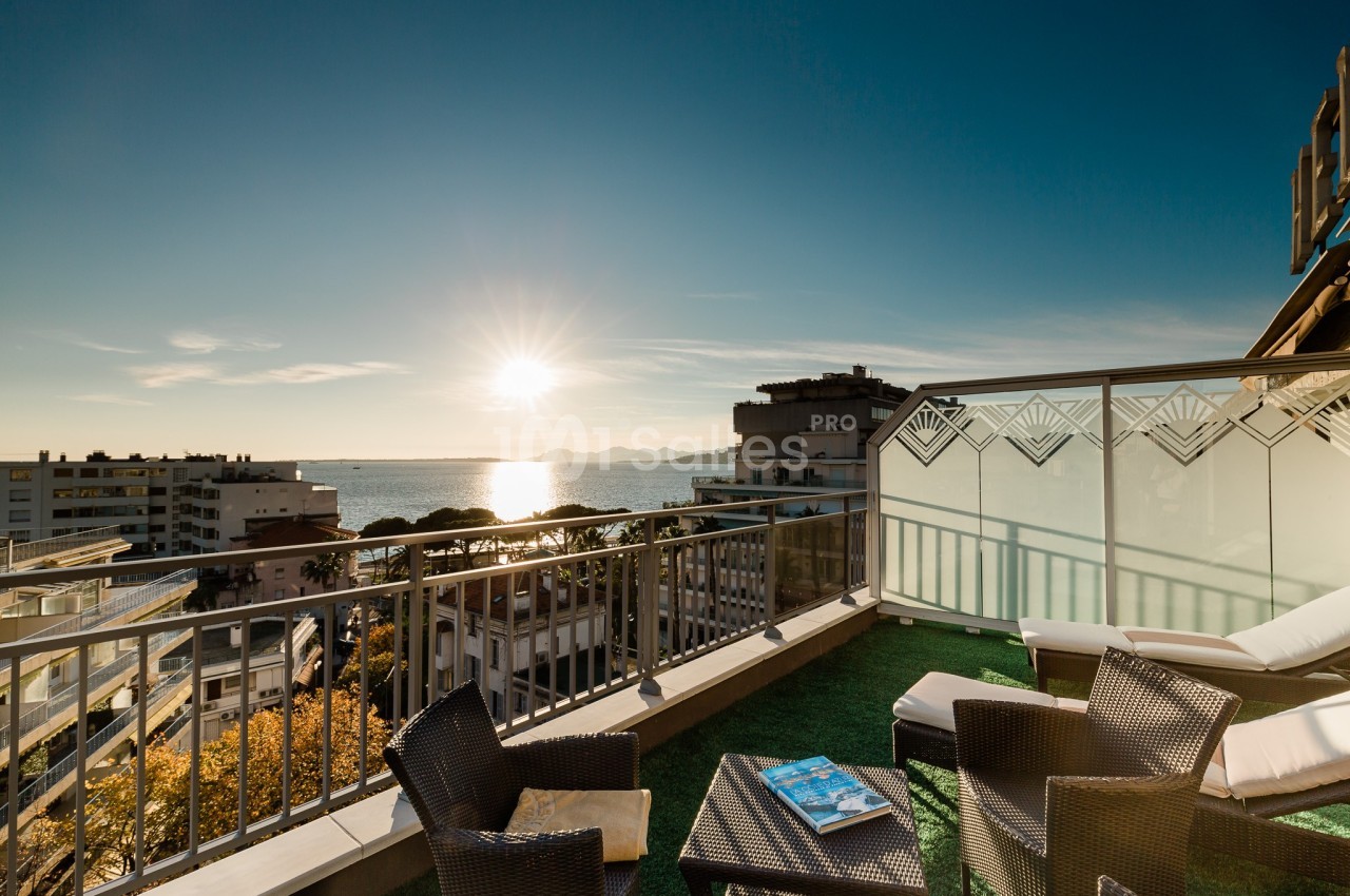 Terrasse avec mobilier en rotin, vue sur la mer au coucher du soleil et bâtiments en arrière-plan.