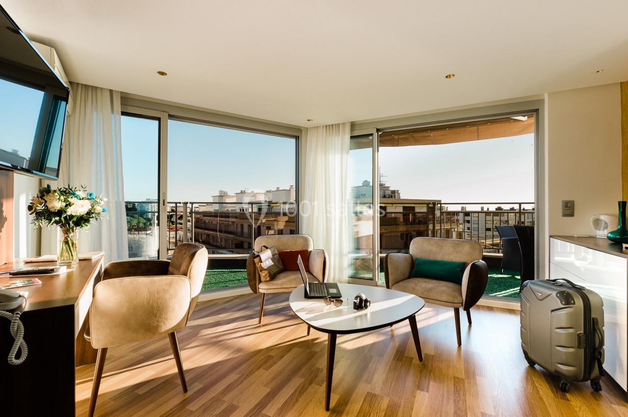 Salon lumineux avec grandes baies vitrées, mobilier moderne, table basse, valise et vue sur des bâtiments urbains.