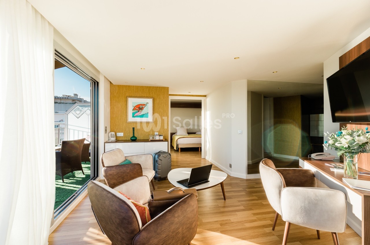 Salon lumineux avec parquet, fauteuils, bureau, télévision, accès à une terrasse et vue sur une chambre au fond.