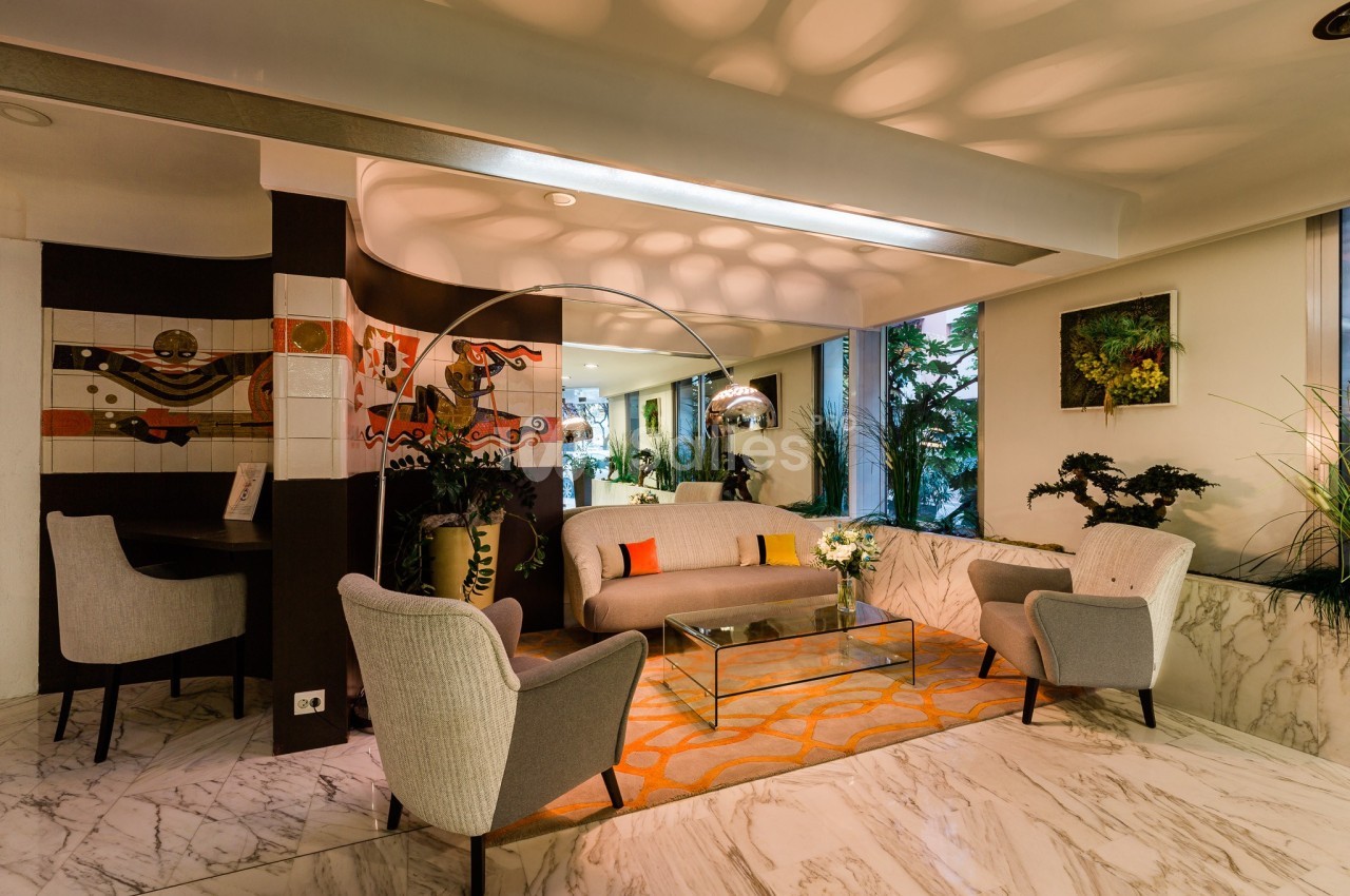 Salon moderne avec canapé, fauteuils gris, table basse en verre, décorations murales colorées et plantes vertes.