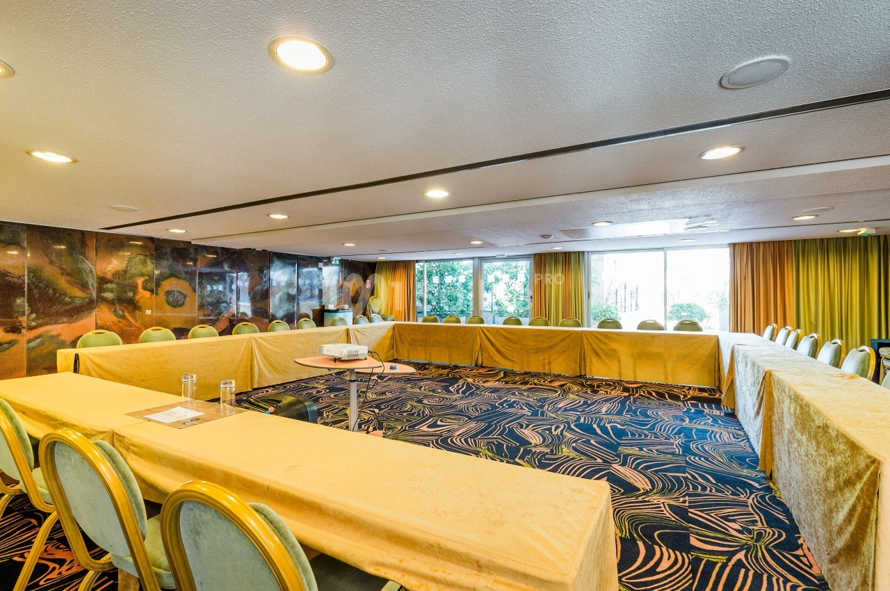 Salle de réunion lumineuse avec tables disposées en U, chaises vertes et grandes fenêtres donnant sur un jardin.