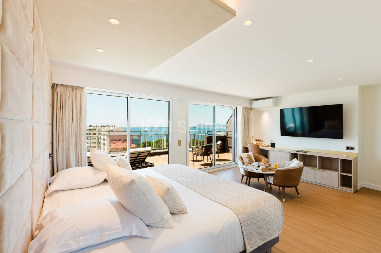 Chambre lumineuse avec lit double, coin salon, télévision murale et terrasse offrant une vue sur la mer.