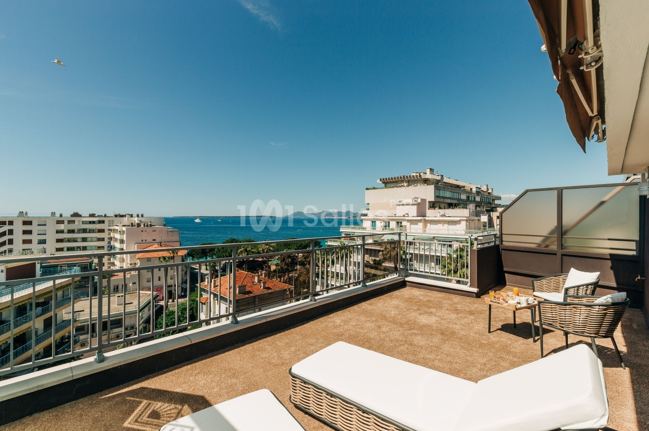 Terrasse ensoleillée avec transats, vue sur la mer, des immeubles et des montagnes à l'horizon.