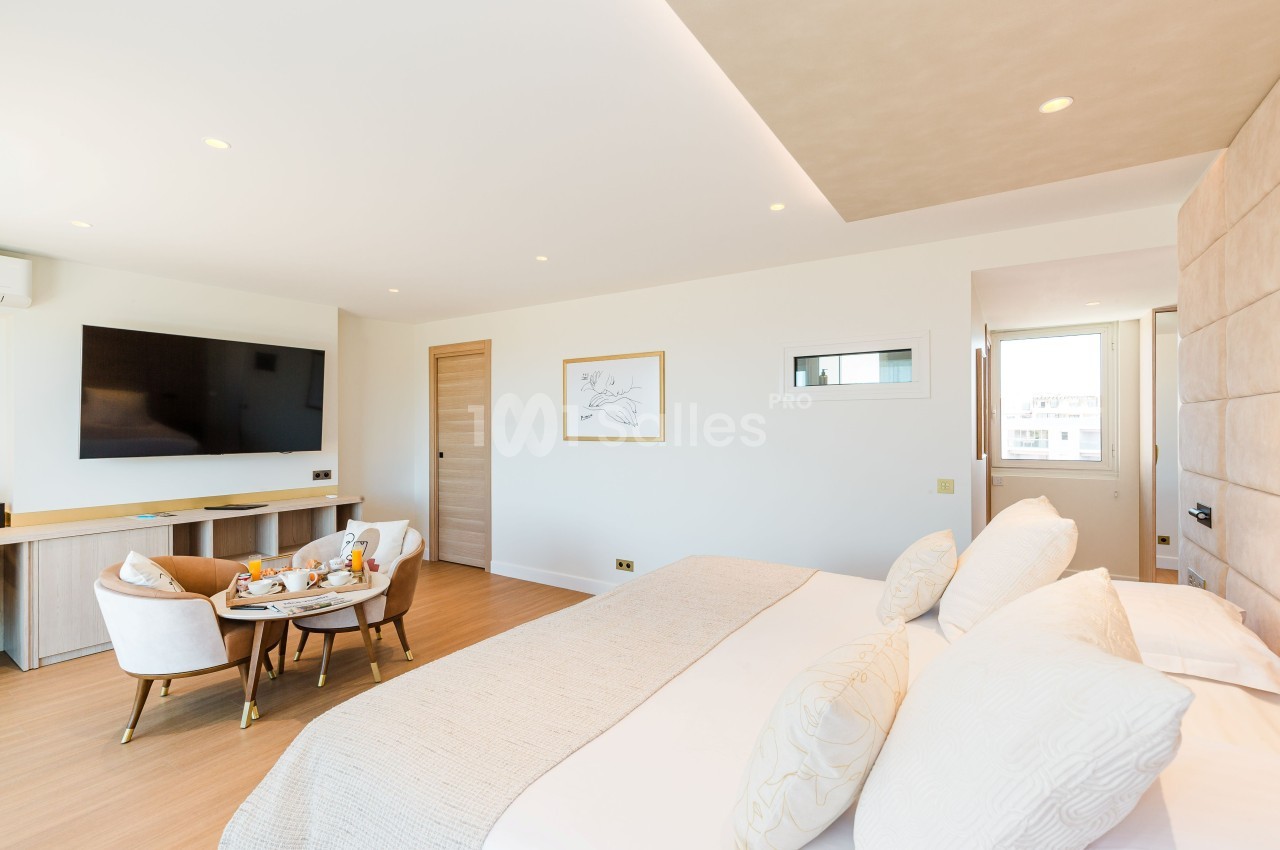 Chambre lumineuse avec lit double, coin salon, télévision murale et table avec plateau-repas, décor moderne et épuré.