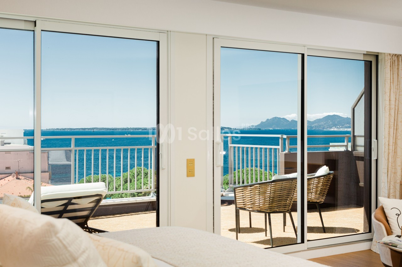 Vue depuis une chambre lumineuse avec des baies vitrées donnant sur une terrasse, la mer et des montagnes au loin.