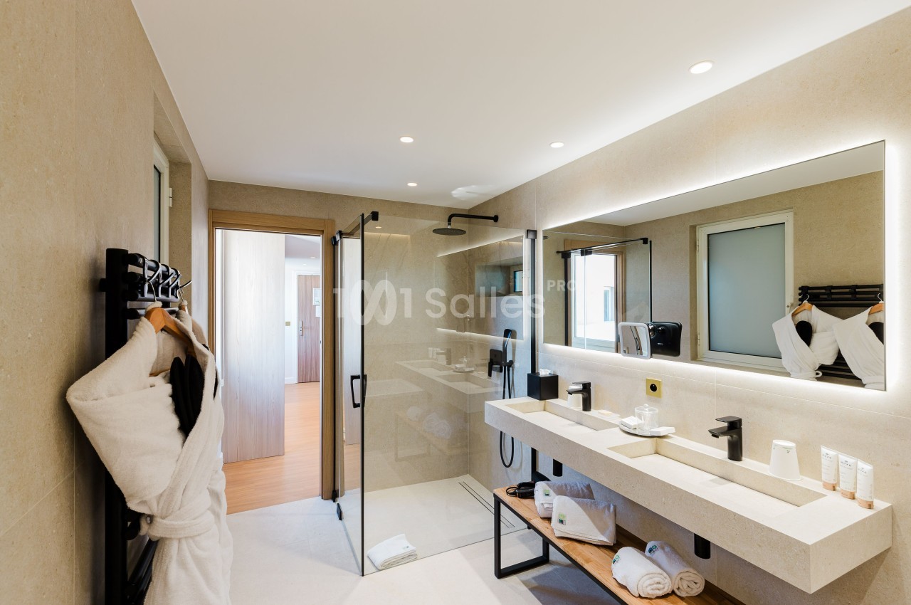 Salle de bain moderne avec douche vitrée, double vasque, miroir éclairé et peignoirs accrochés.