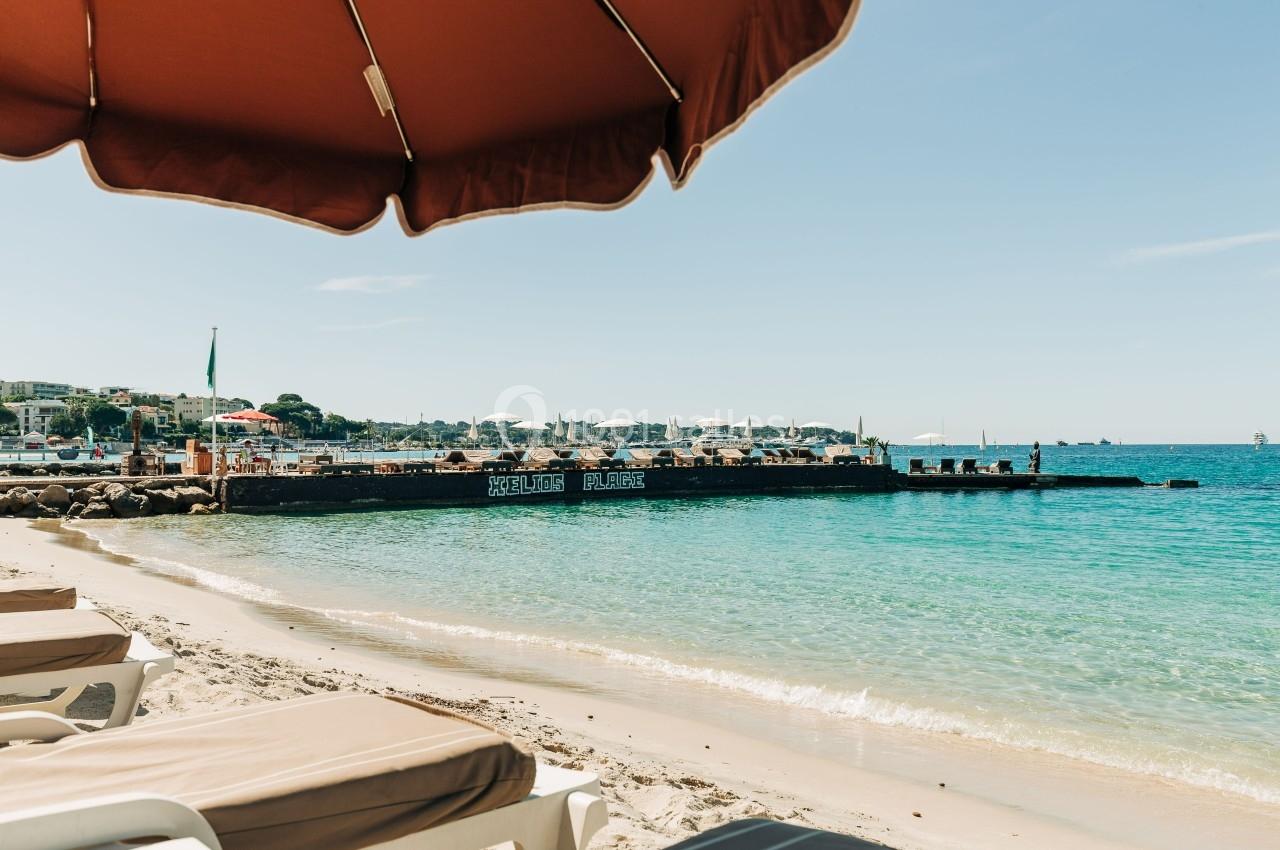 Plage avec transats sous un parasol, eau turquoise et ponton avec parasols en arrière-plan.