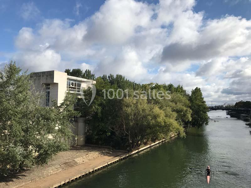 Vue d'une rivière bordée d'arbres avec un bâtiment à gauche et une personne pagayant sur l'eau.