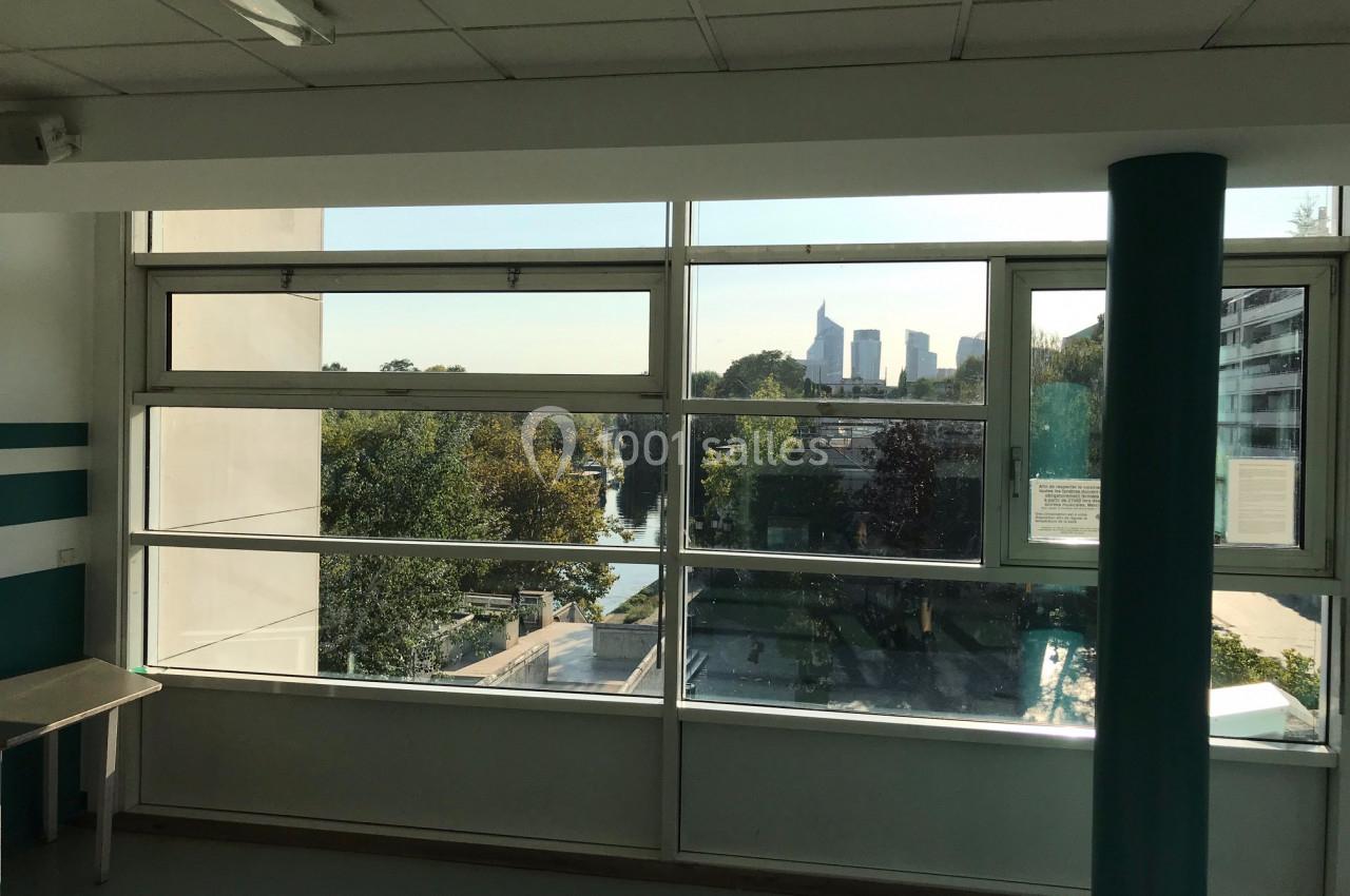 Vue depuis une fenêtre d'intérieur sur un paysage urbain avec des arbres, un fleuve et des immeubles en arrière-plan.
