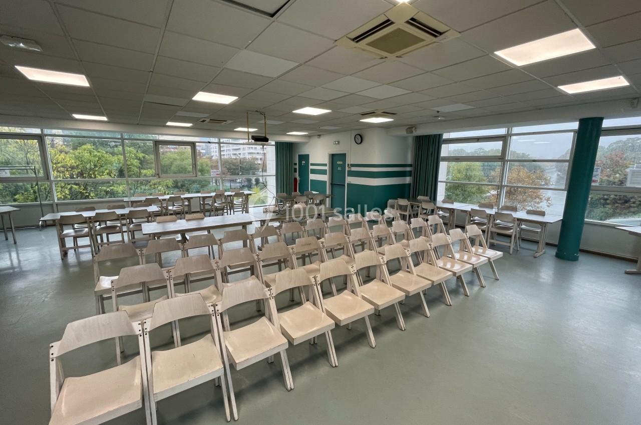 Salle lumineuse avec des rangées de chaises blanches alignées, grandes fenêtres donnant sur des arbres et bâtiments.