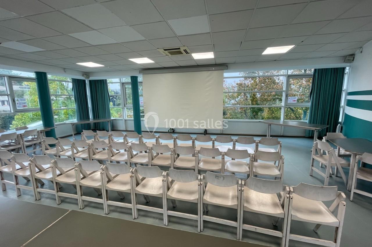 Salle de conférence lumineuse avec des rangées de chaises blanches alignées face à un écran de projection.