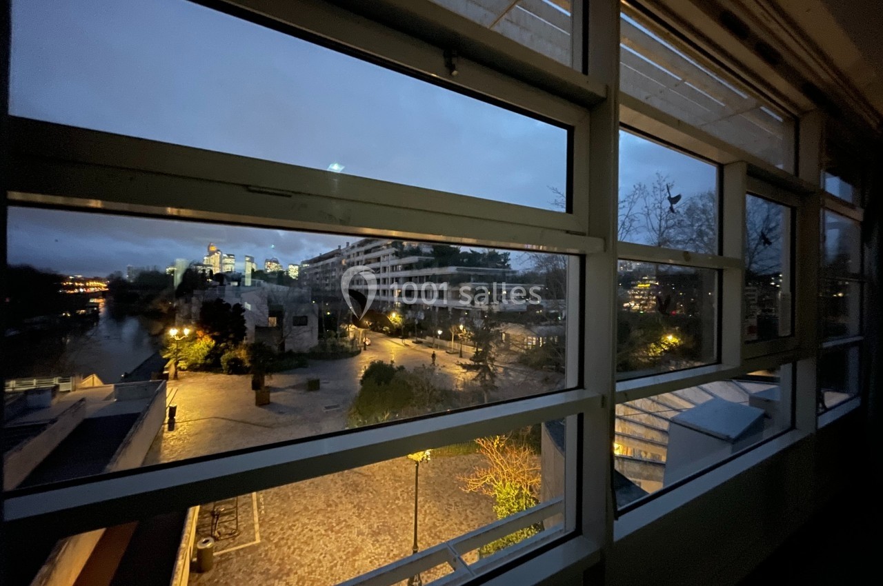 Vue nocturne d'une place éclairée avec des bâtiments modernes et un cours d'eau, observée depuis une fenêtre.