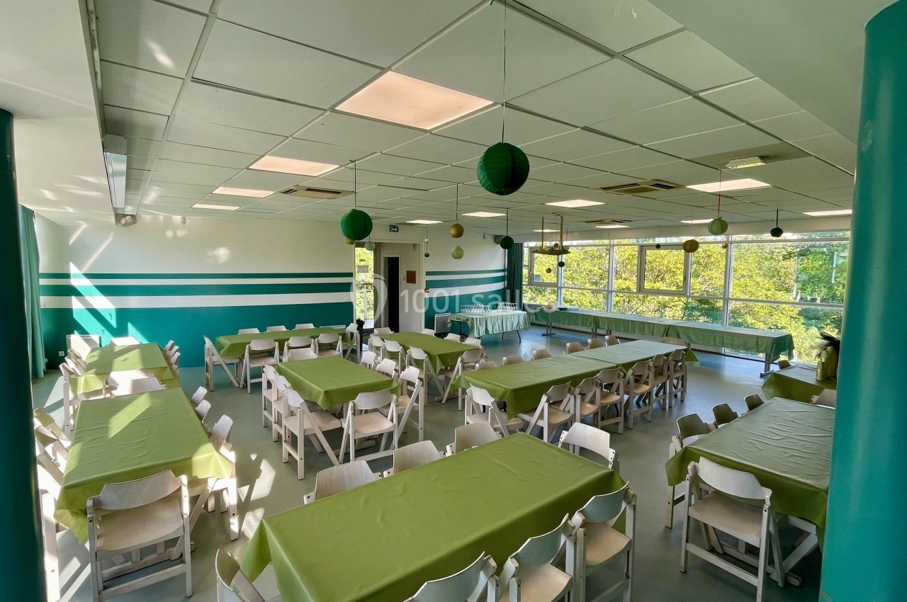 Salle lumineuse avec des tables recouvertes de nappes vertes, chaises blanches et décorations suspendues.