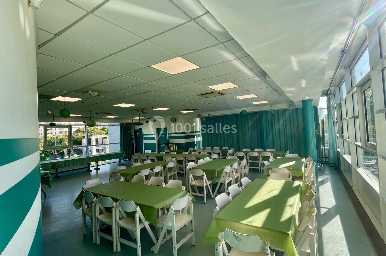 Salle lumineuse avec des tables recouvertes de nappes vertes, entourées de chaises blanches, et de grandes fenêtres.