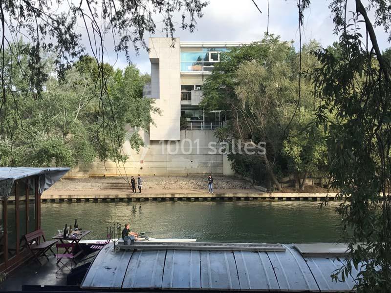 Vue d'un bâtiment moderne entouré d'arbres, situé au bord d'un canal avec une péniche et un rameur au premier plan.