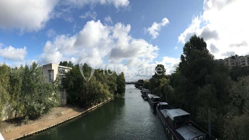 Vue d'un canal bordé d'arbres avec des péniches amarrées, sous un ciel partiellement nuageux.