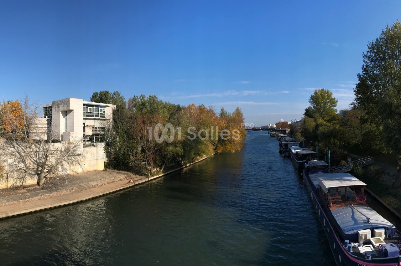 Vue d'un canal bordé d'arbres en automne, avec des péniches amarrées et un bâtiment moderne sur la rive gauche.