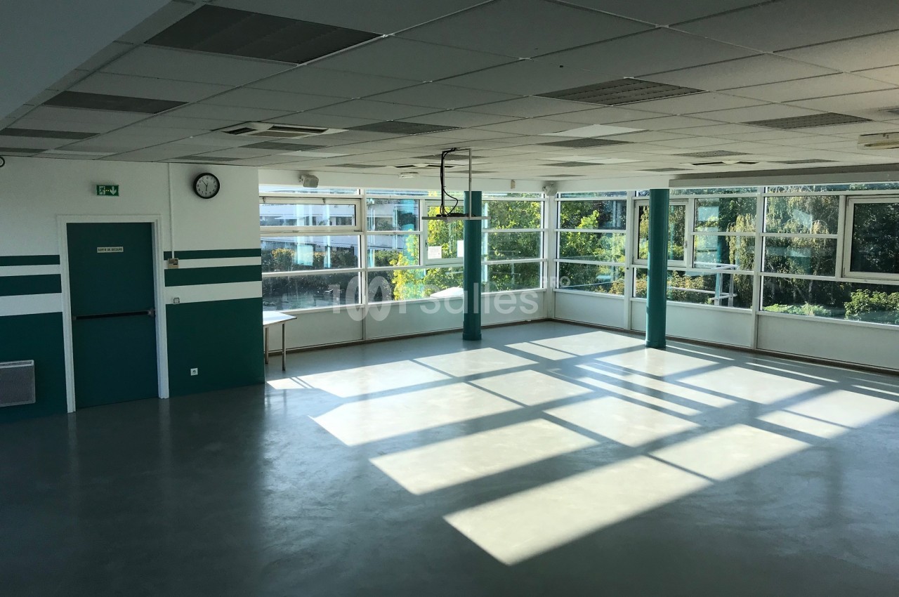 Salle lumineuse avec de grandes fenêtres, murs blancs et verts, sol clair, et vue sur des arbres à l'extérieur.