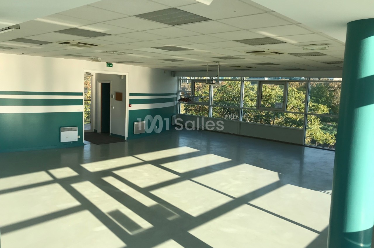 Salle vide avec sol en béton, murs blancs et verts, grandes fenêtres laissant entrer la lumière naturelle.