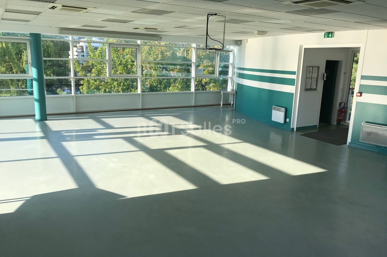 Salle vide avec sol en béton, murs blancs et verts, grandes fenêtres laissant entrer la lumière naturelle.