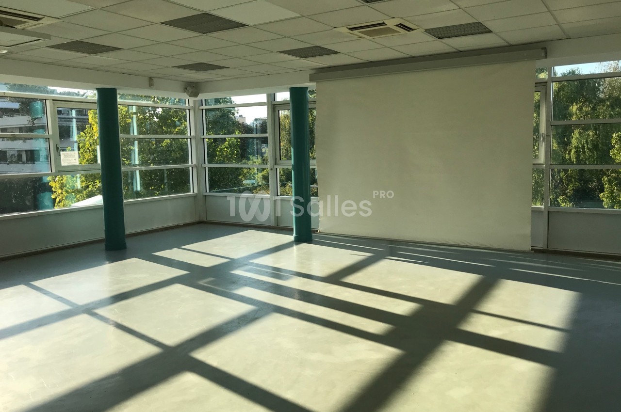 Salle vide avec un sol en béton, des murs vitrés laissant entrer la lumière naturelle et un écran blanc au fond.