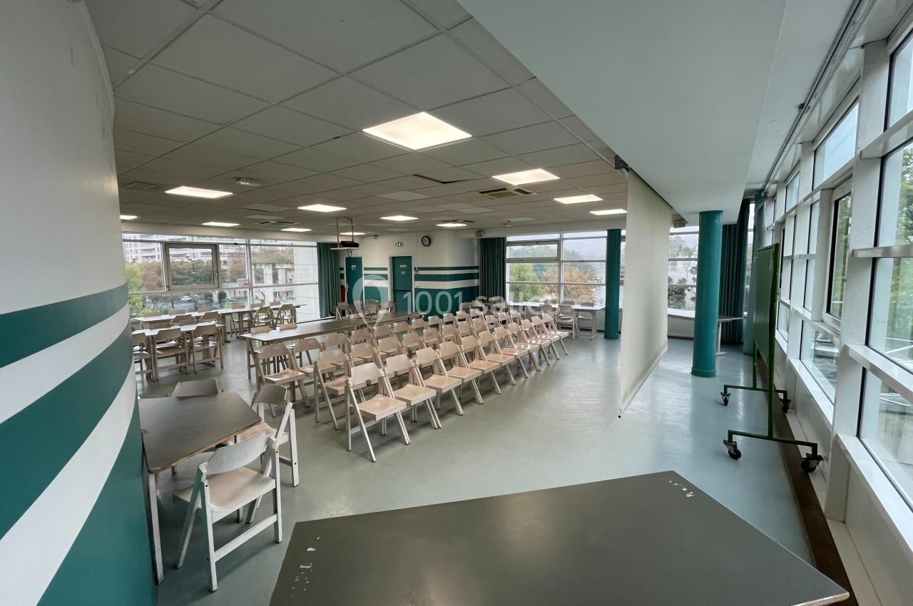 Salle lumineuse avec des rangées de chaises blanches, tables et un écran de projection, entourée de grandes fenêtres.