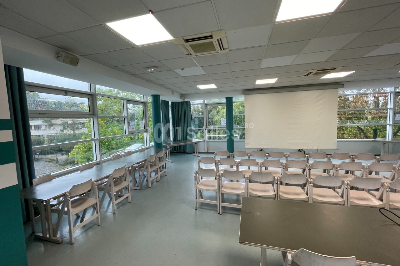 Salle de conférence lumineuse avec des rangées de chaises, une grande baie vitrée et un écran de projection.