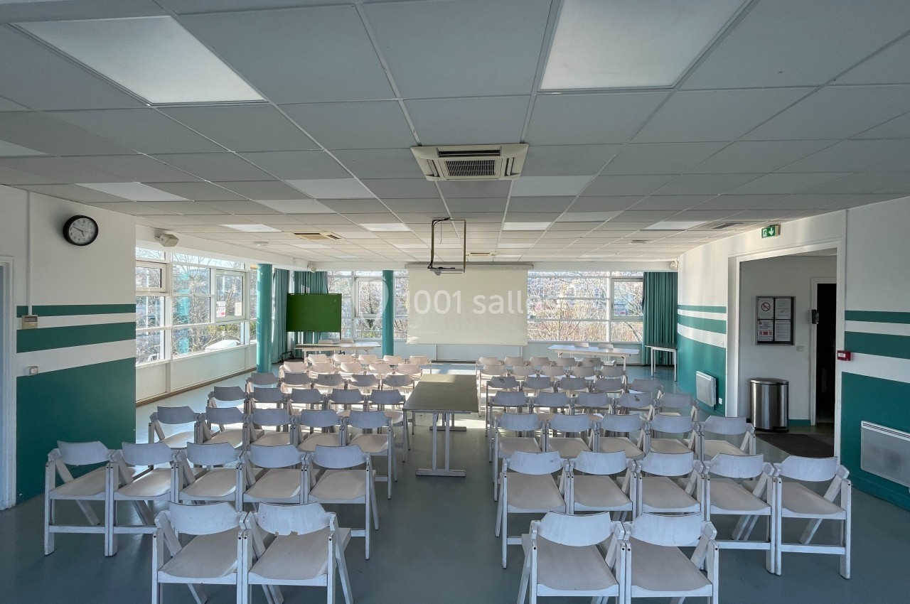 Salle de conférence lumineuse avec des rangées de chaises blanches, un écran de projection et de grandes fenêtres.