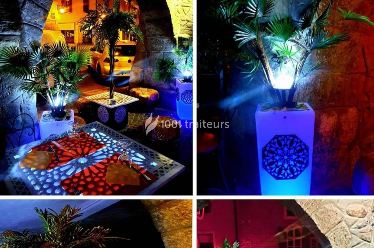 Terrasse éclairée de nuit avec tables décorées, plantes en pot et ambiance lumineuse colorée sous des arches en pierre.