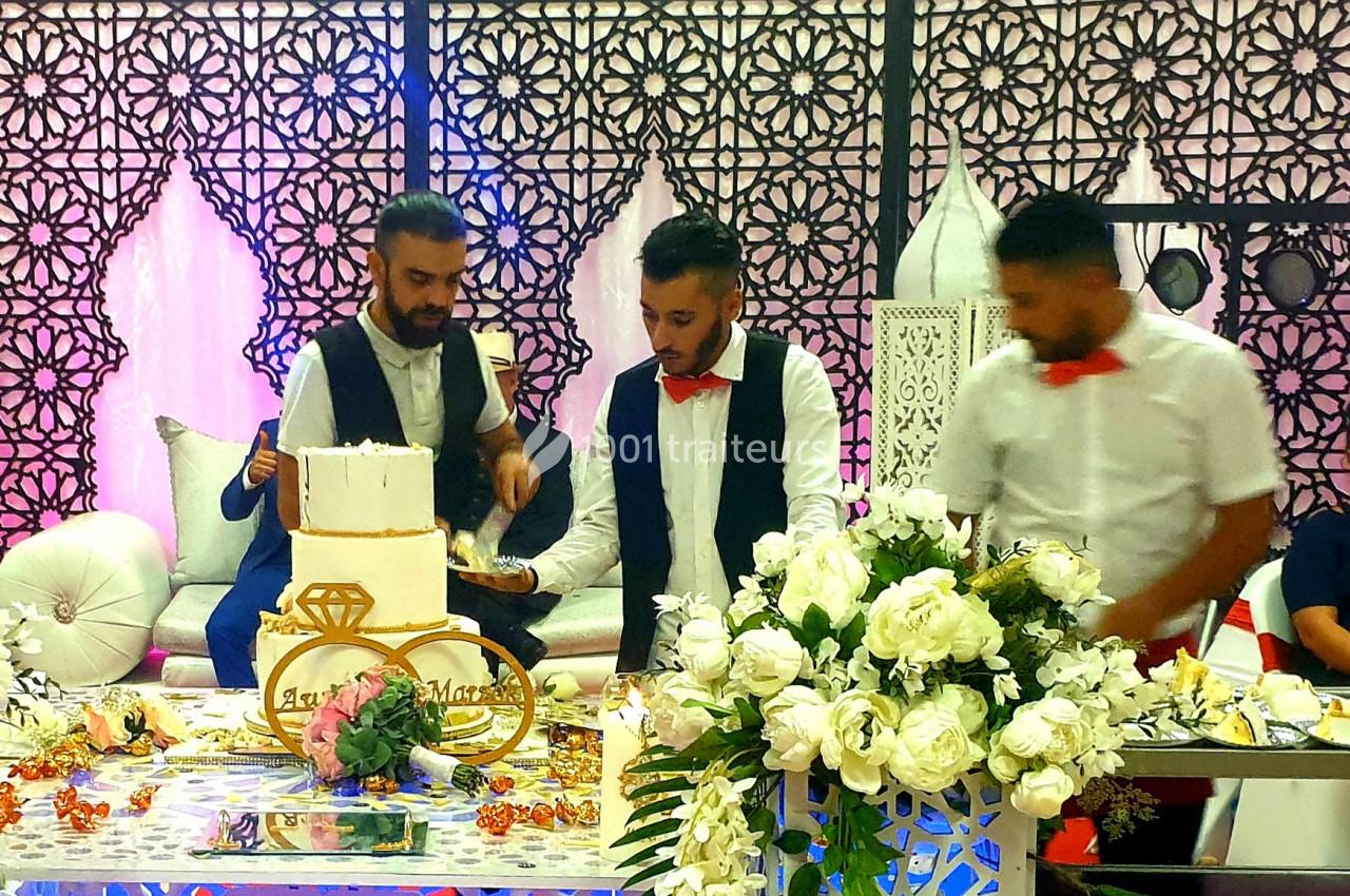 Trois hommes en tenue de cérémonie décorent et préparent un gâteau de mariage devant une table ornée de fleurs blanches.