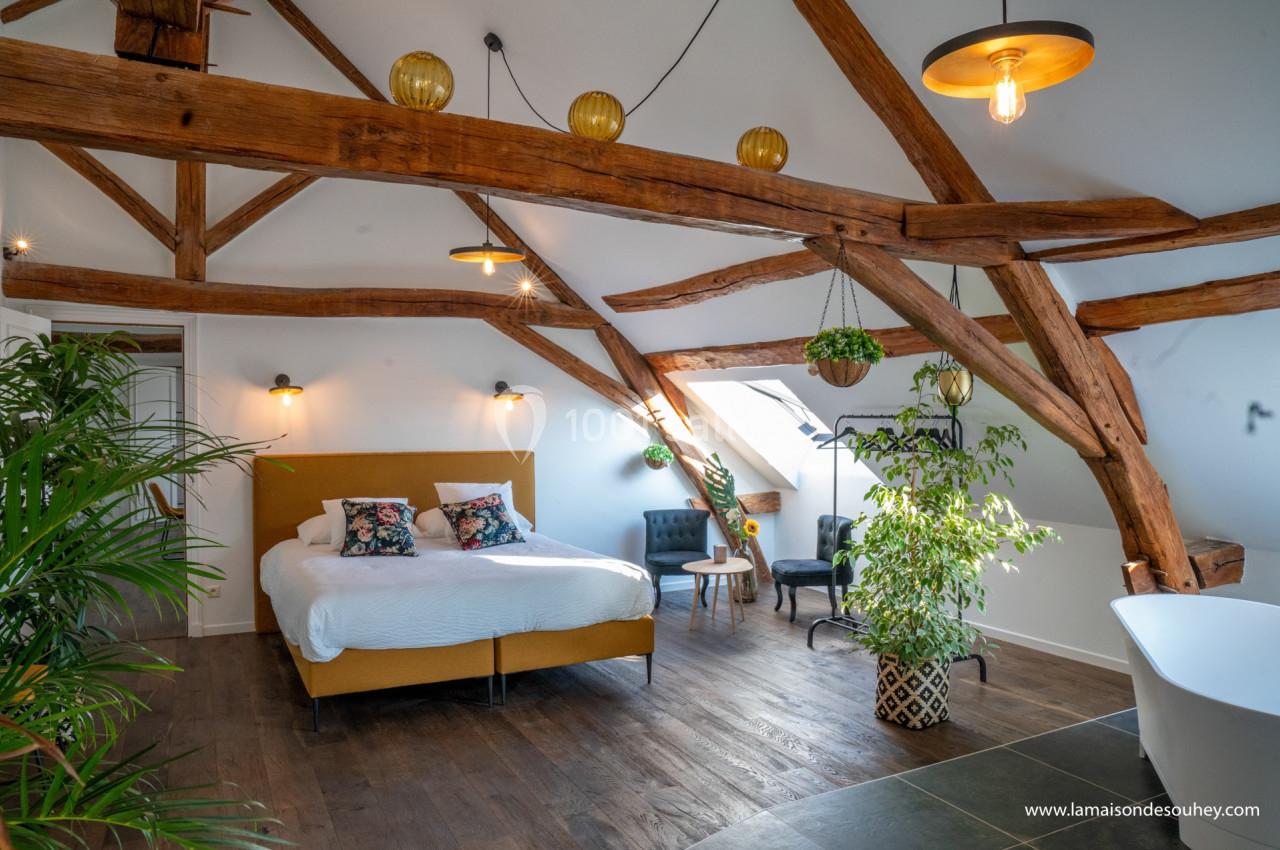 Chambre lumineuse sous combles avec poutres apparentes, lit double, plantes, fauteuils et baignoire.