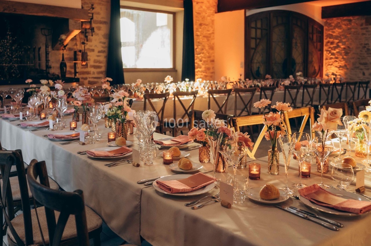 Salle de réception décorée avec des tables alignées, nappes blanches, fleurs pastel et bougies allumées.