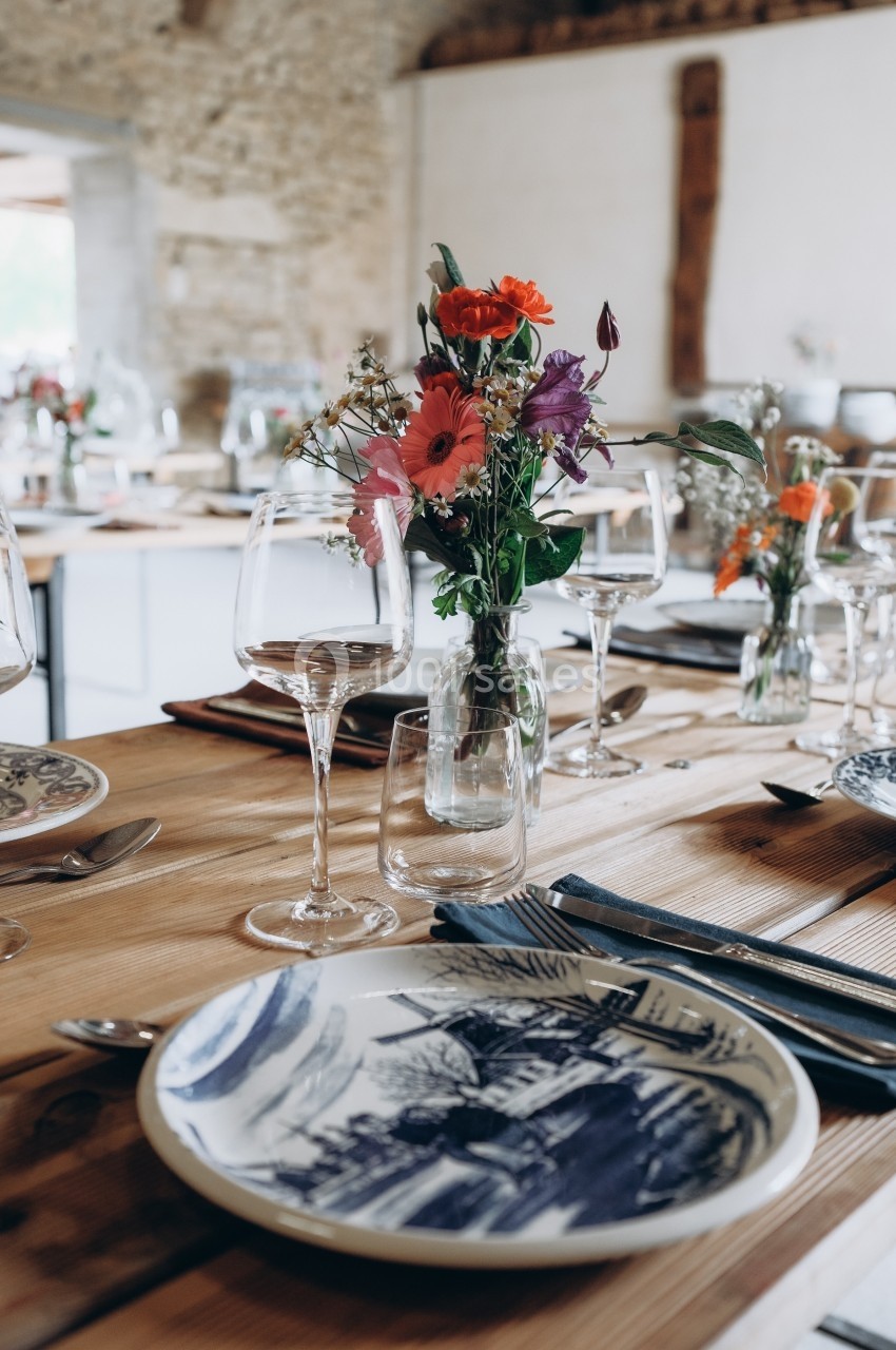 Table en bois dressée avec des assiettes décorées, verres à vin et un bouquet de fleurs colorées au centre.