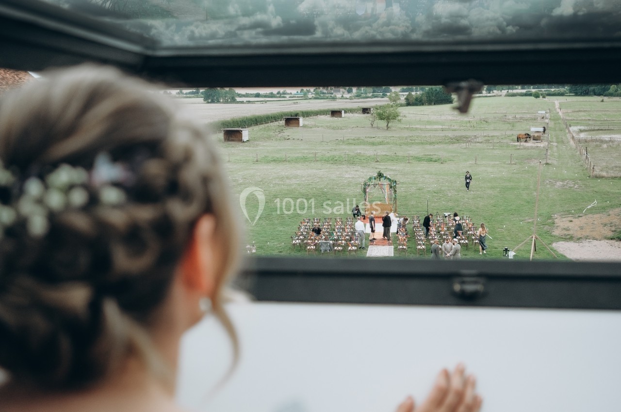 Vue depuis une fenêtre sur une cérémonie de mariage en plein air avec des invités assis et une arche décorée.