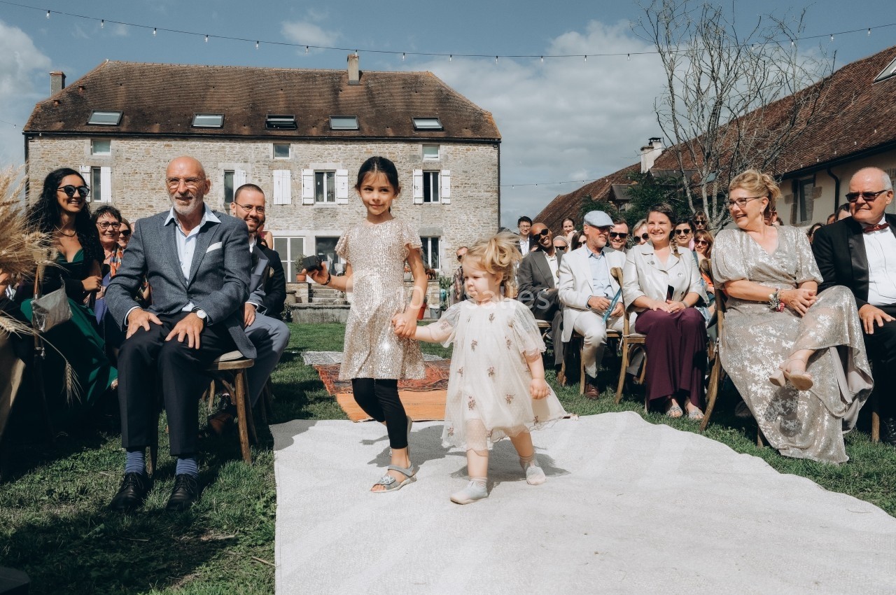 Deux enfants marchent main dans la main sur une allée blanche lors d'une cérémonie en plein air, entourés d'invités…