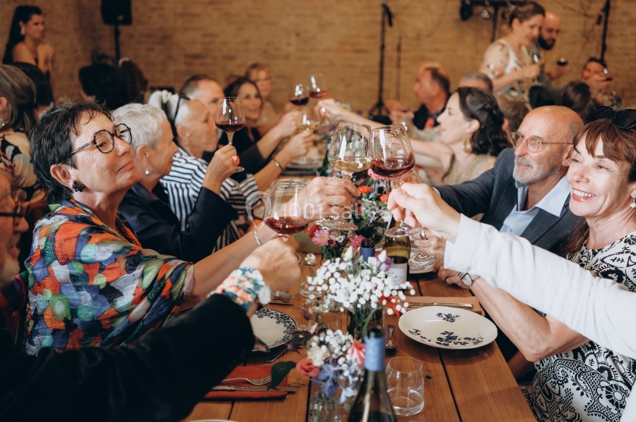 Des convives trinquent avec des verres de vin autour d'une grande table lors d'un repas convivial en intérieur.