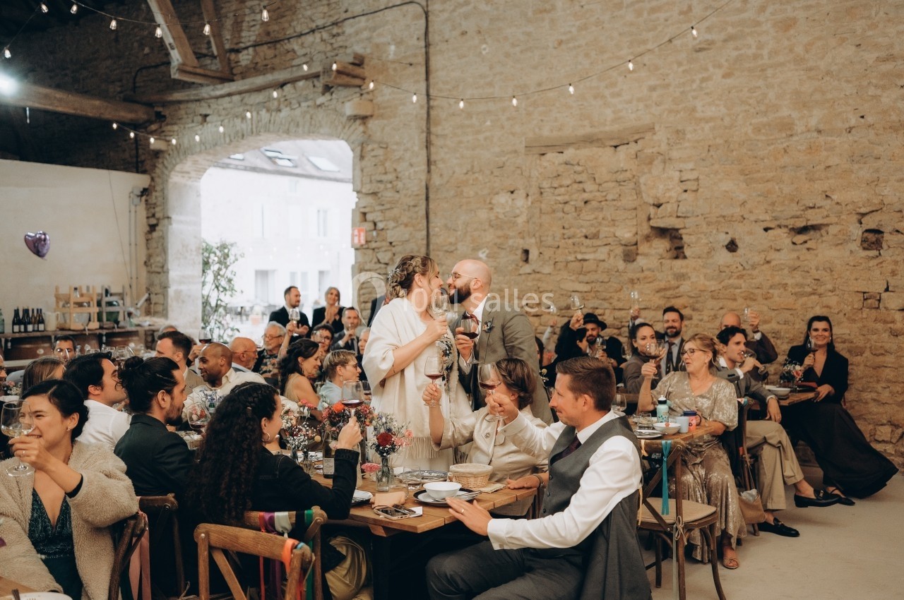 Un couple échange un baiser lors d'un repas de mariage dans une salle en pierre, entouré d'invités souriants.