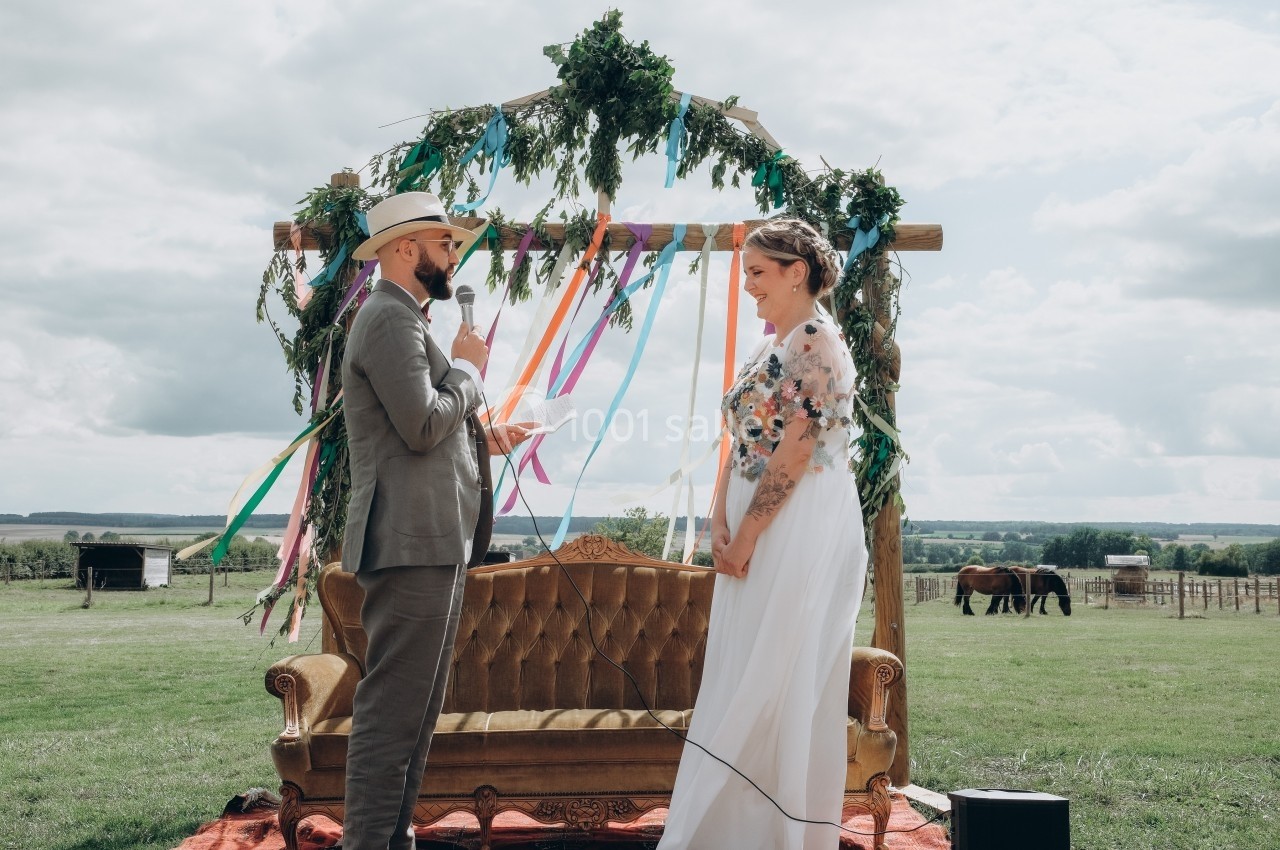 Un couple échange ses vœux devant une arche décorée de rubans colorés dans un cadre champêtre.