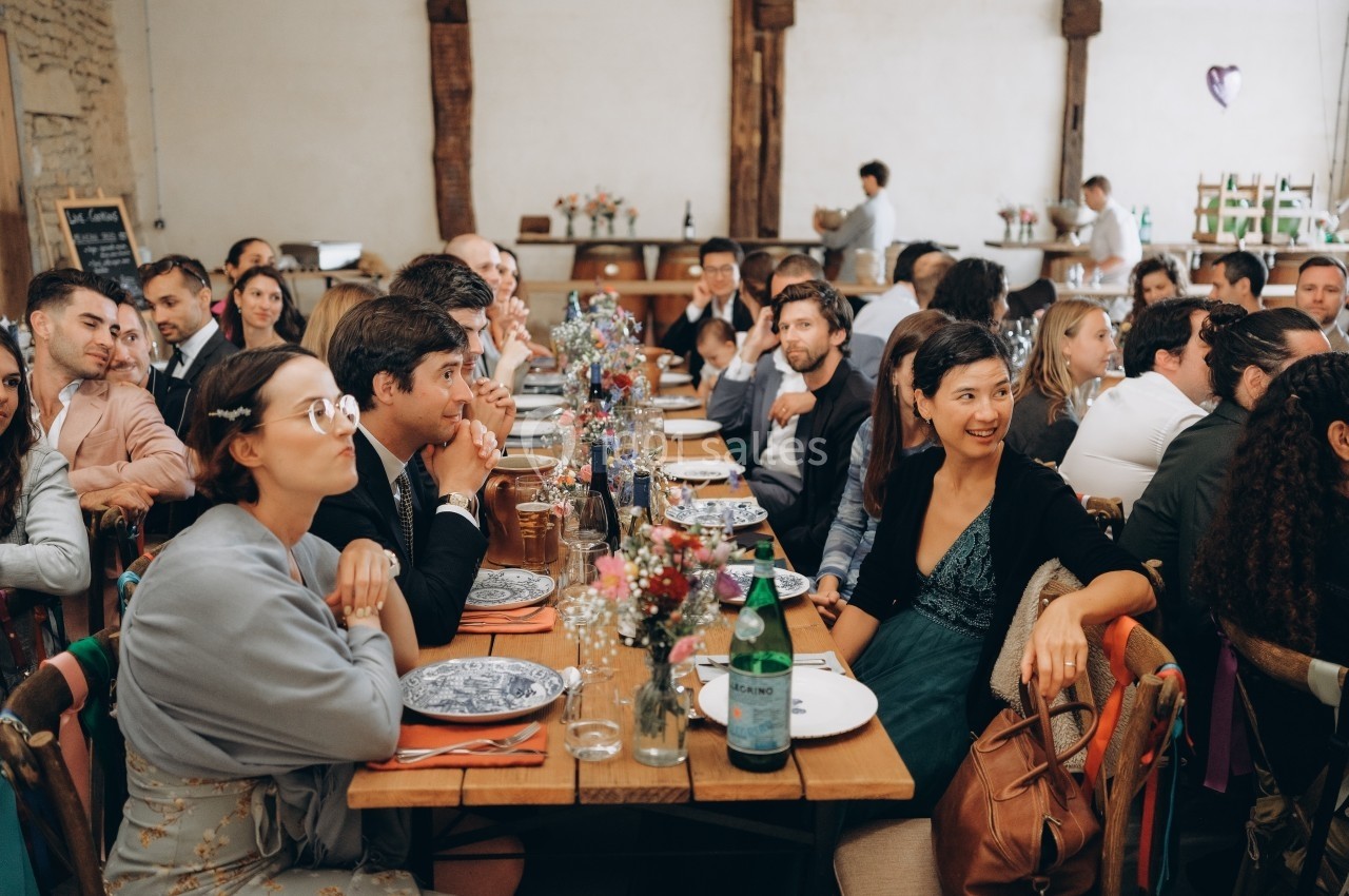Des convives assis à une longue table décorée, lors d'un repas dans une salle au style rustique.
