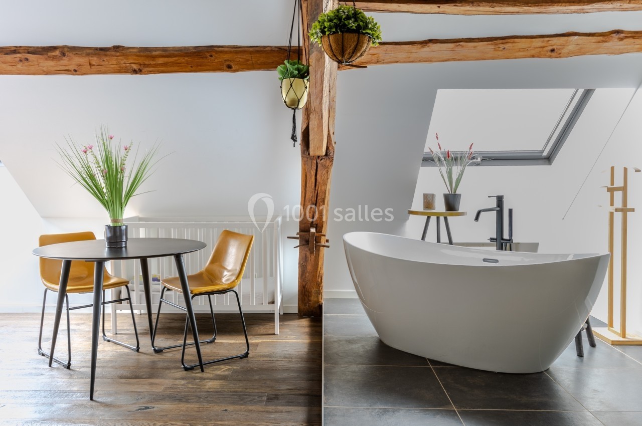 Salle de bain lumineuse avec baignoire îlot, table ronde, chaises en cuir et poutres apparentes sous un toit mansardé.