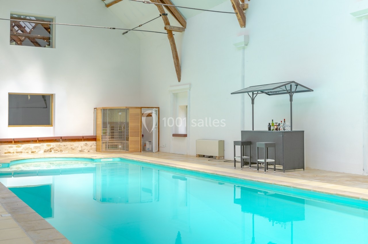 Piscine intérieure avec bar aménagé et cabine en bois, entourée de murs blancs et éclairée par la lumière naturelle.