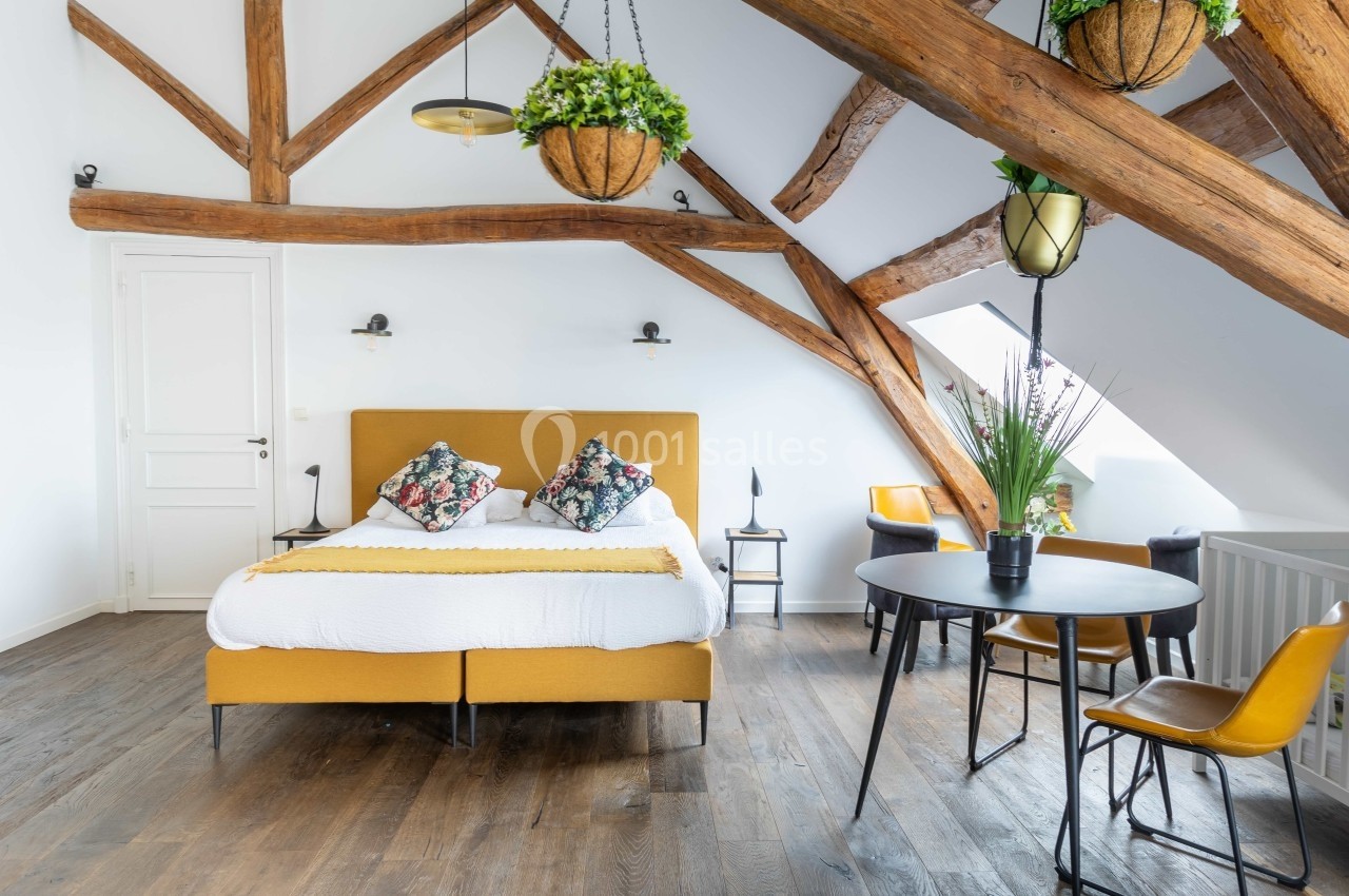Chambre mansardée lumineuse avec poutres apparentes, lit double, table ronde, chaises et plantes suspendues.