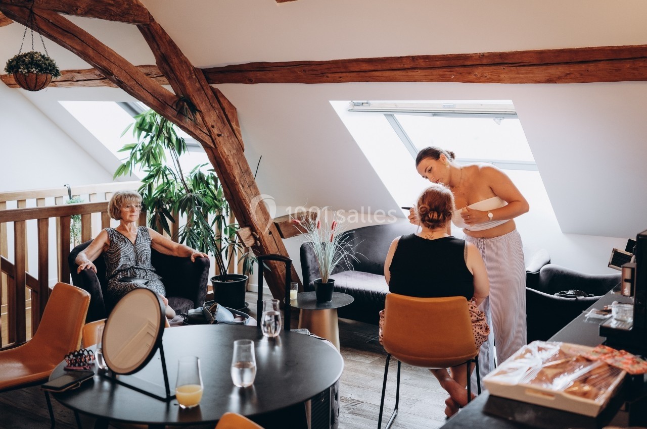 Deux femmes discutent pendant une séance de maquillage dans un salon lumineux avec poutres apparentes, une autre femme…