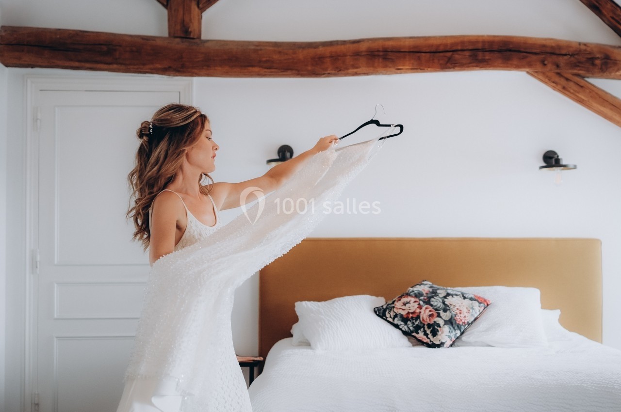 Femme tenant une robe blanche devant un lit dans une chambre lumineuse avec poutres apparentes.