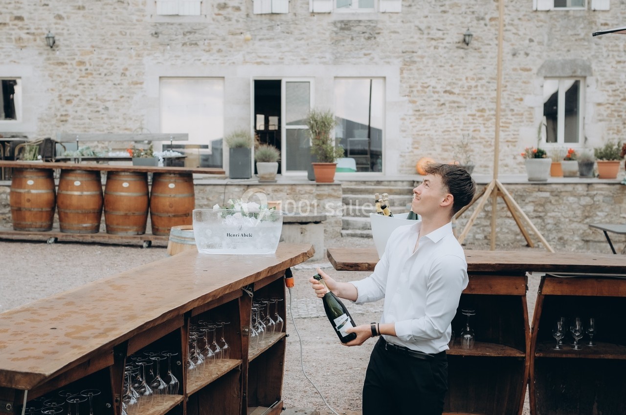 Un homme en chemise blanche ouvre une bouteille de champagne derrière un bar extérieur devant un bâtiment en pierre.